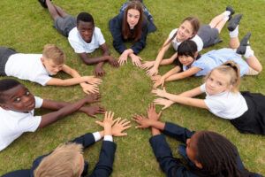 group of school children laying on grass in a circle, like a wheel, with hands crossed in front of them